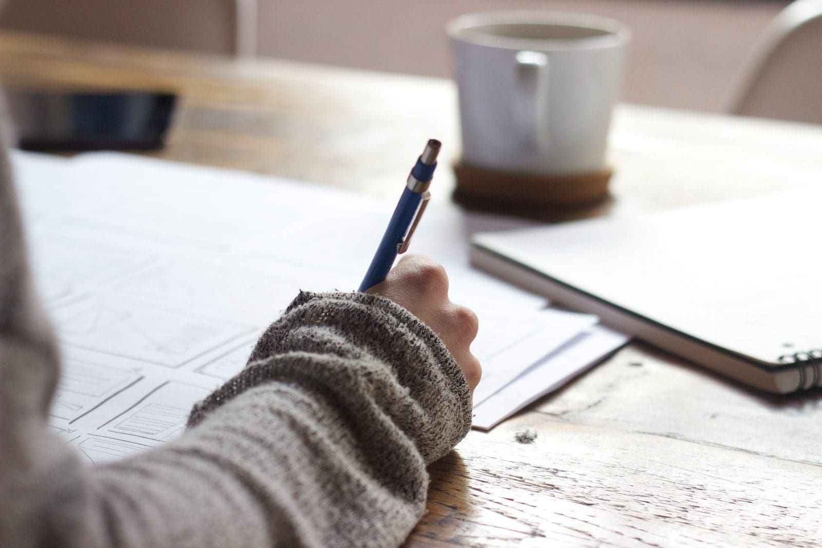 person writing on brown wooden table near white ceramic mug college essy